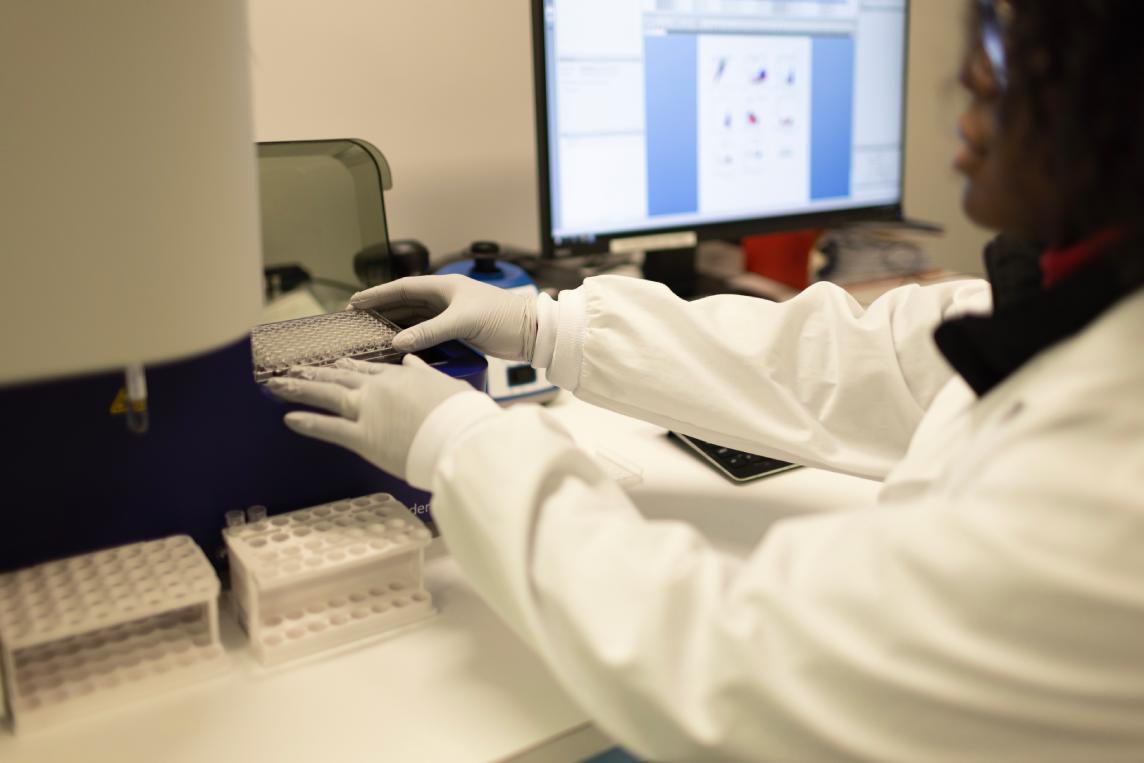 Scientist loading samples into a rack beside a monitor displaying flow cytometry plots.